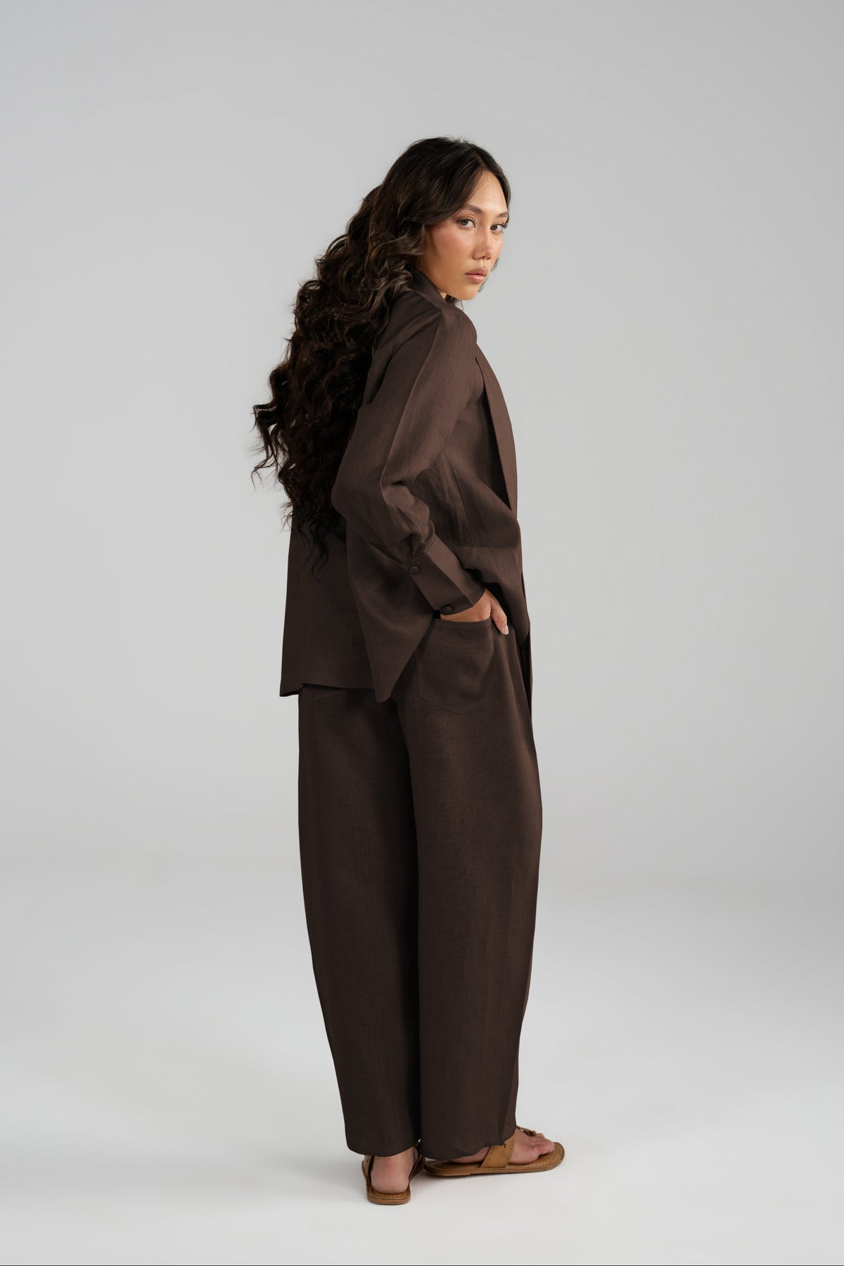 A woman modeling a dark brown two-piece outfit consisting of a long-sleeve blazer and matching wide-leg trousers, standing against a light gray studio background.