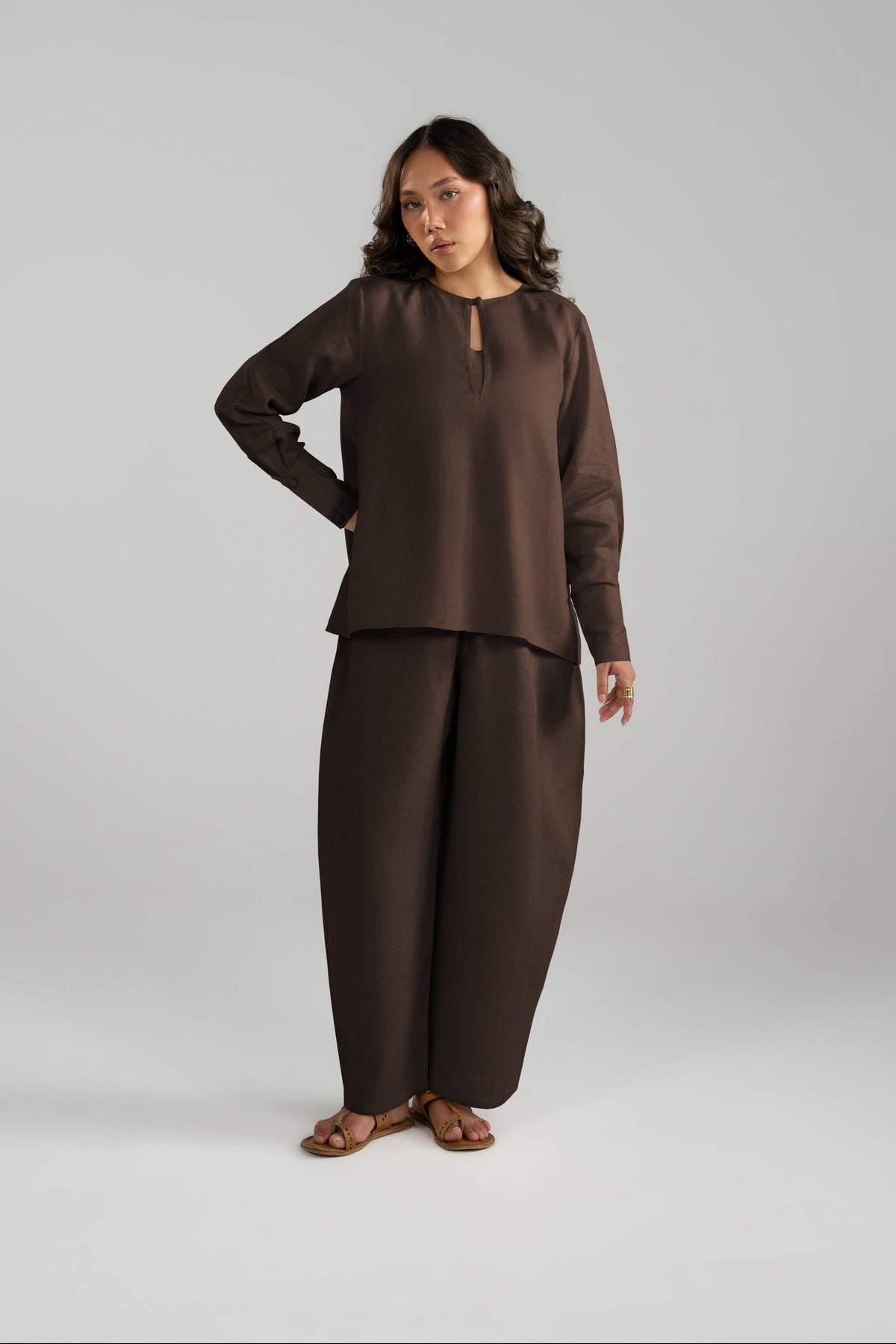 A woman modeling a dark brown two-piece linen outfit consisting of a long-sleeve top with a keyhole neckline and matching wide-leg pants.
