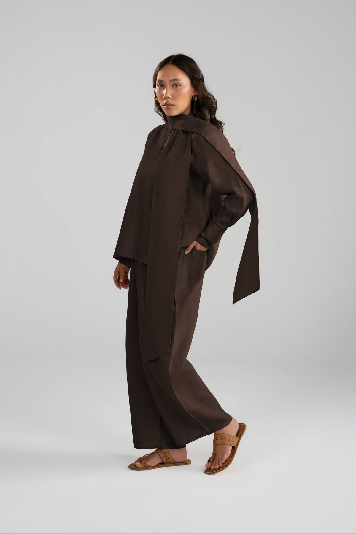 A model wearing a dark brown, minimalist two-piece outfit consisting of a draped long-sleeve top and matching wide-leg trousers, paired with brown leather sandals.