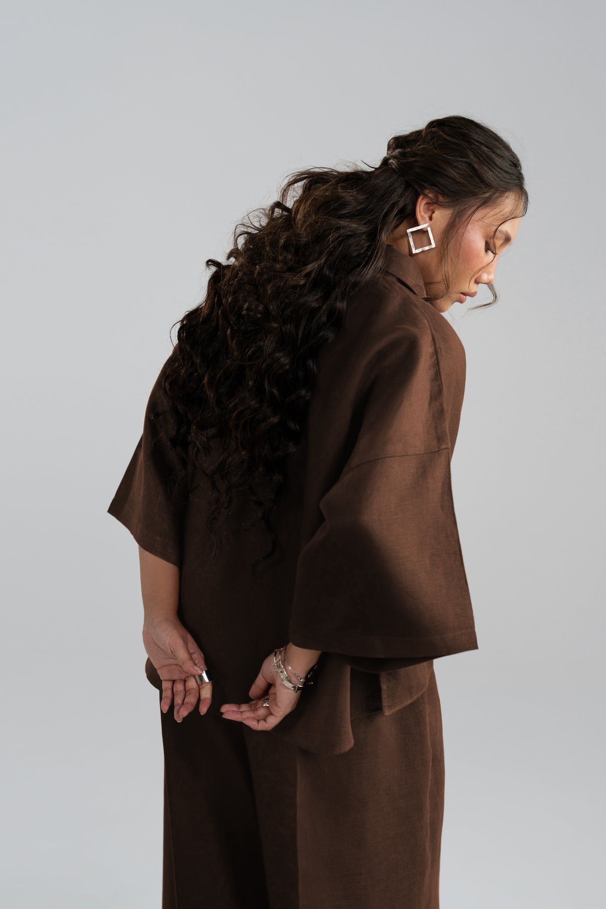 Woman wearing an espresso brown matching set with a wide draped sleeve, viewed from the side with hands behind her back.