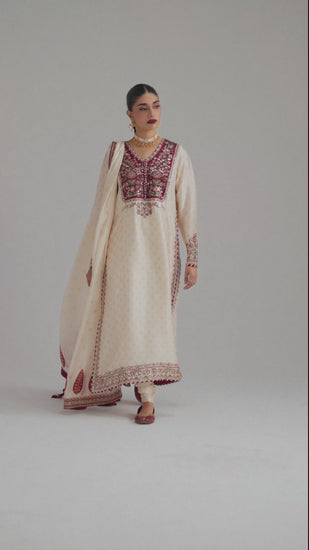 The Dove Set by Mannam is an ivory printed long kameez with a distinctive red applique neckline, resham and sequin embroidery, chatta patti trim on the neck, sleeves and hem, and contrast maroon facings with embroidered buttons. It comes with a classic churidaar and a printed silk dupatta with goti and tassels. 