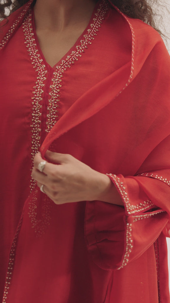 The Flame Set by Mannam is a pure silk kameez in burnt orange featuring a V-neckline with organza trimmings, artisanal hand-stitched embroidery along the neckline, sleeves, side seams and hem, floral embroidery at the hem and distinctive cut-out sleeve detailing. It comes with pure silk trousers and a pure organza dupatta with hand embroidery.