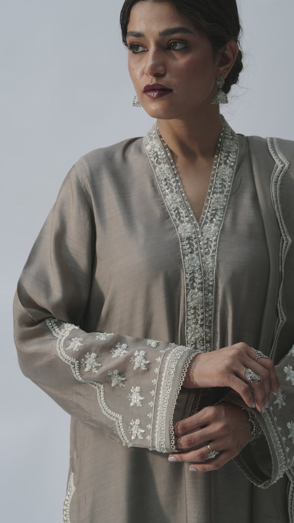 The Mink Set by Mannam is a pure silk V-neck kameez in a warm mink tone, featuring hand-embellished sequins on the neckline, hem and sleeves, organza finishing on the sleeves, and floral motifs at the hem. It comes with pure silk embroidered trousers with organza finishing and a pure organza dupatta with a scallop-edged embroidered border and floral motifs.